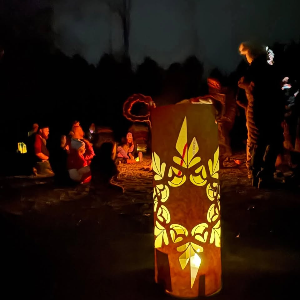 A handmade paper lantern glowing at the Lantern Walk with families gathered around a fire in the background