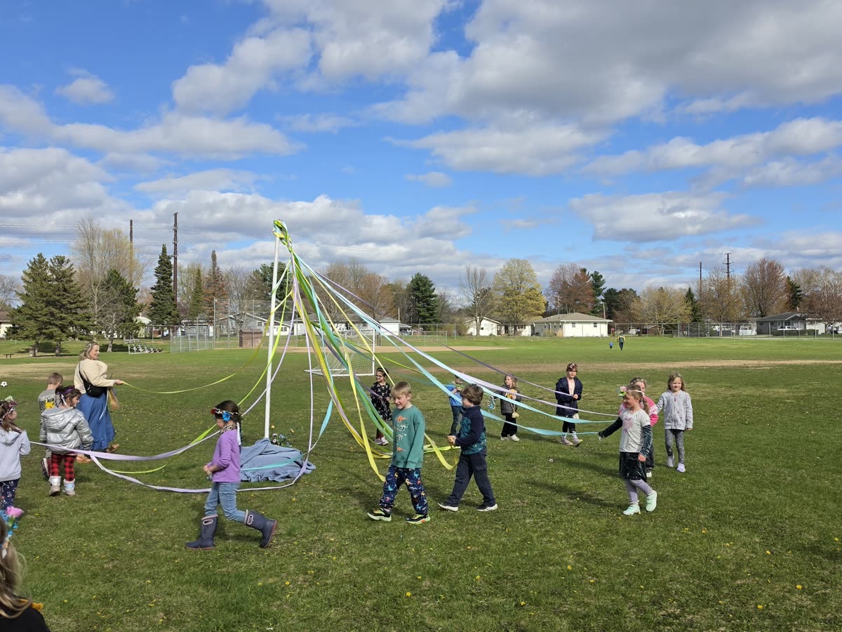 Students winding pastel ribbons around a maypole in the school field under a blue spring sky