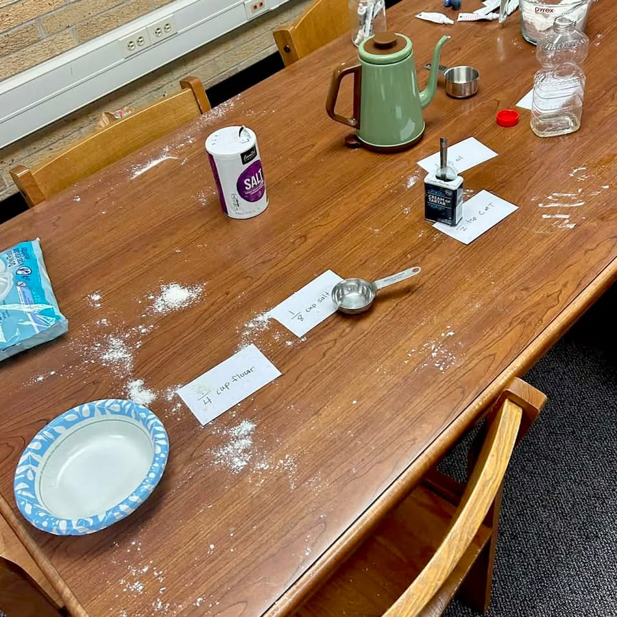 A classroom table set with flour, salt, cream of tartar and a measuring cup, beside handwritten cards reading one quarter cup flour and one eighth cup salt