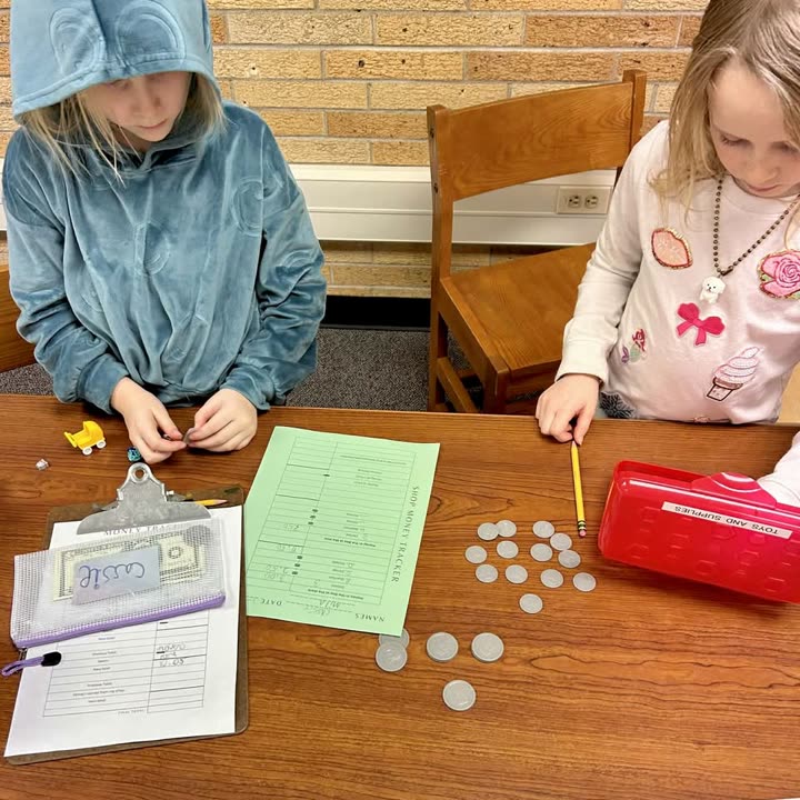Two students working with coins, worksheets, and a cash box at a classroom table
