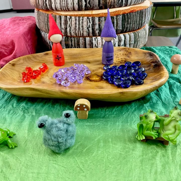 A seasonal nature table with peg dolls, felted frogs, and coloured glass stones in a wooden bowl