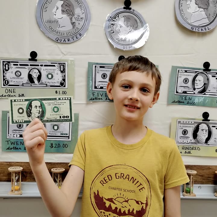 A student in a Red Granite Charter School shirt holding a paper bill in front of a classroom display of coins and currency