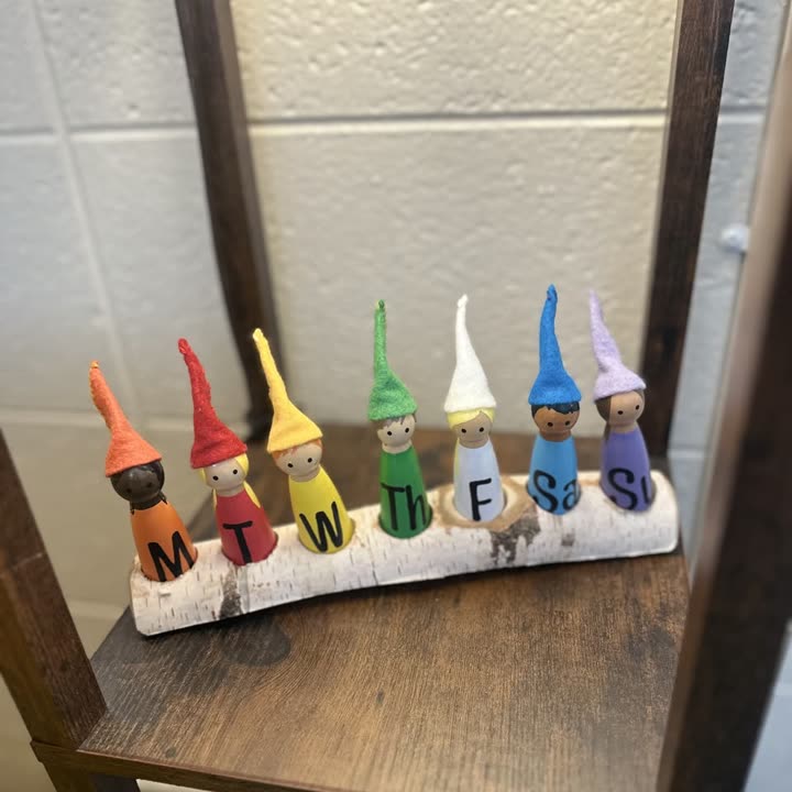 Seven peg dolls in coloured felt hats labelled with the days of the week, standing on a birch log